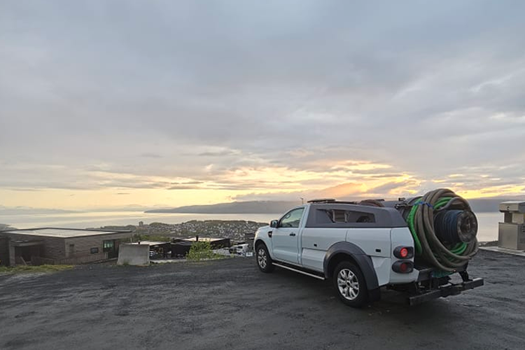 En hvit pickup med slange står på en vei med utsikt over havet og solnedgang i bakgrunnen.