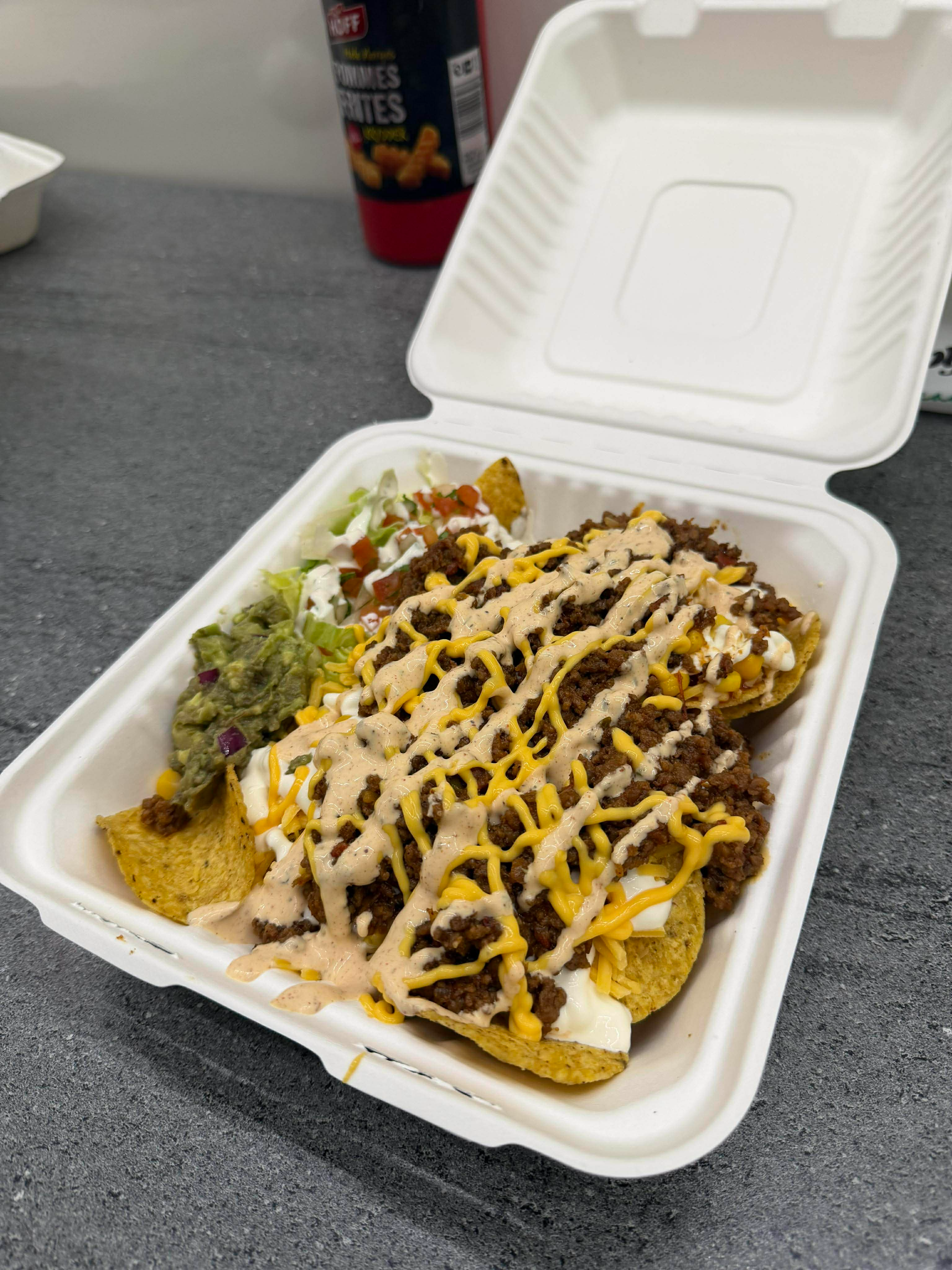 En take-away-boks med nachos toppet med kjøttdeig, ost, guacamole, salat og salsa.