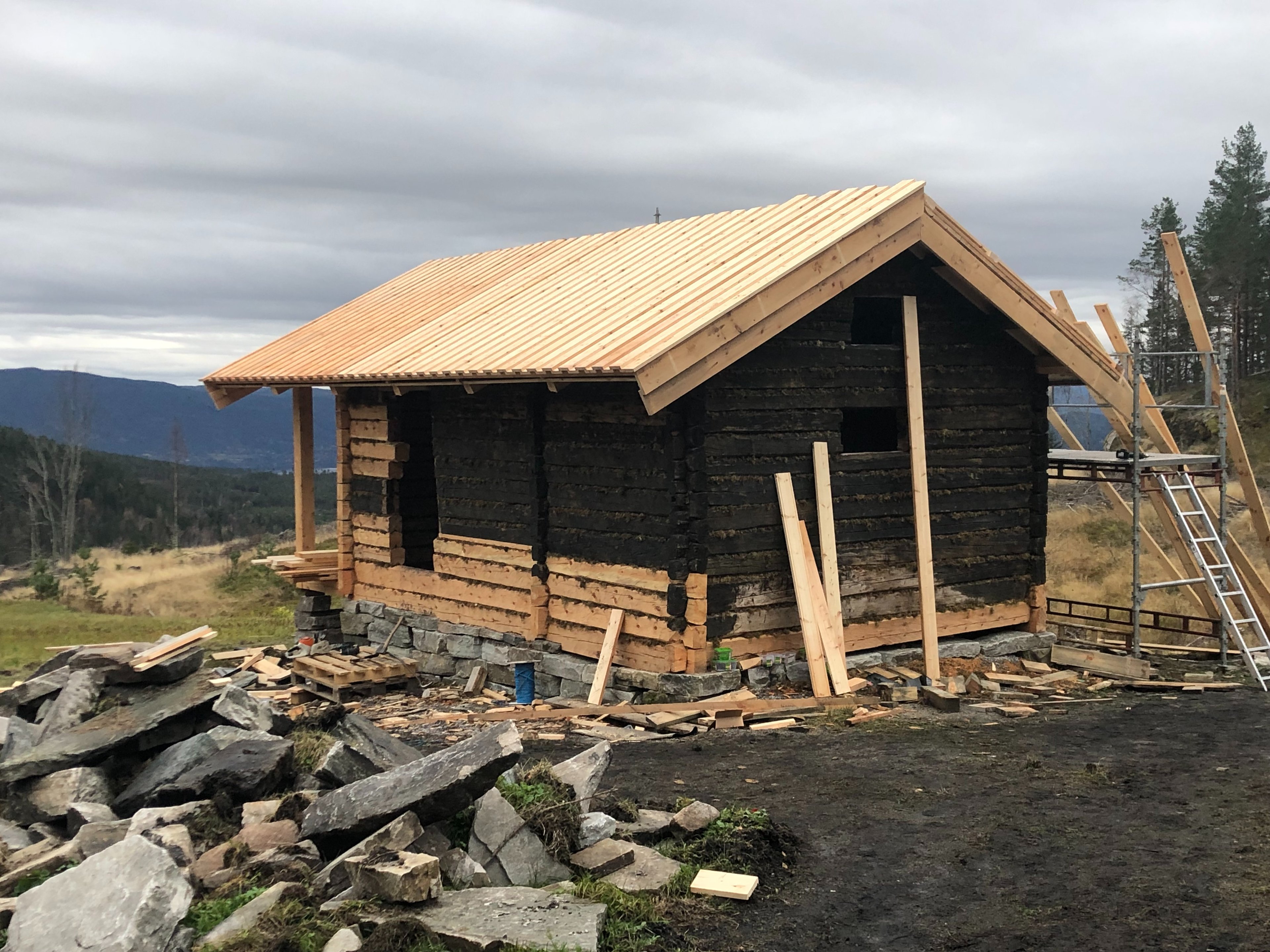 Et påbygg av en hytte med treverk og takplater, omringet av stein og byggeutstyr i et naturskjønt landskap.