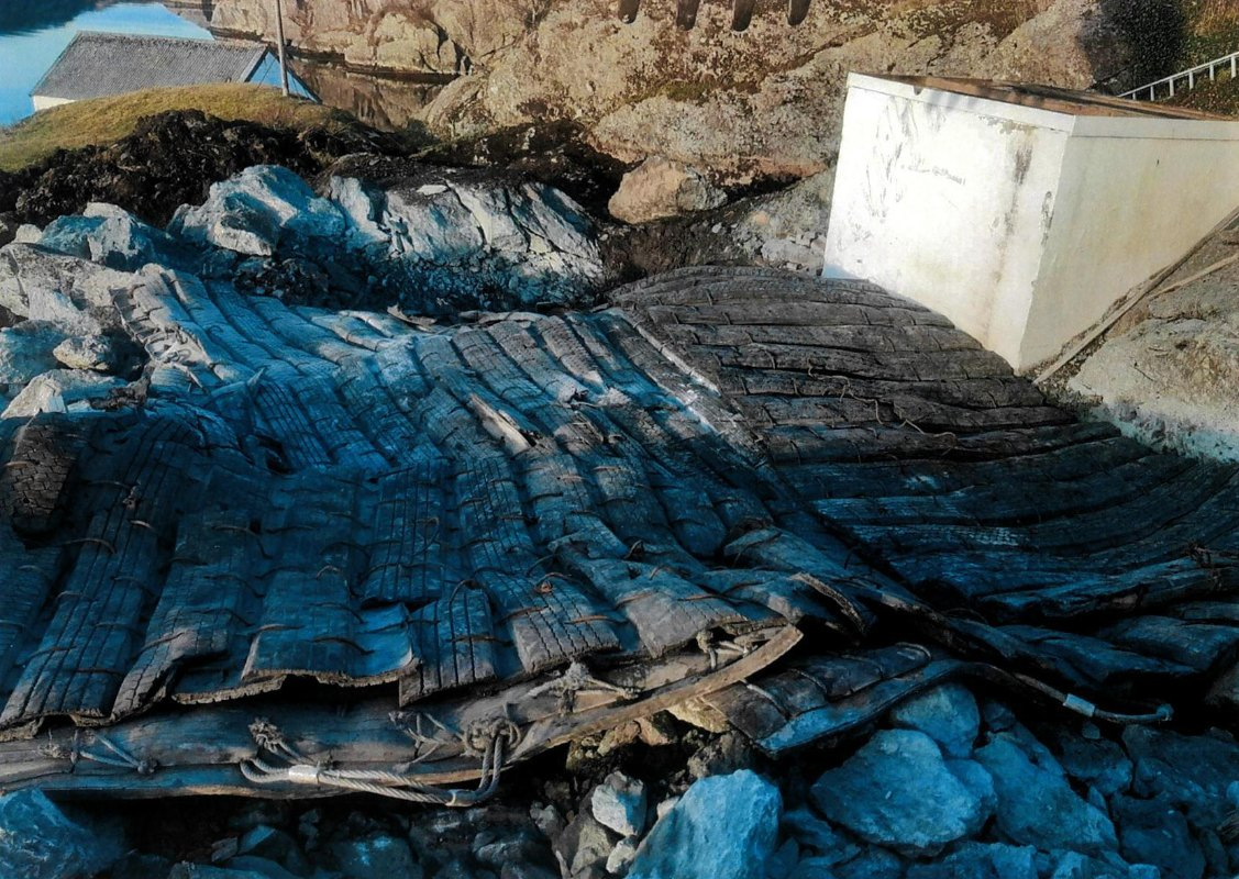 En ødelagt trebro som ligger på steiner ved kysten, med en hvit betongstruktur i bakgrunnen.