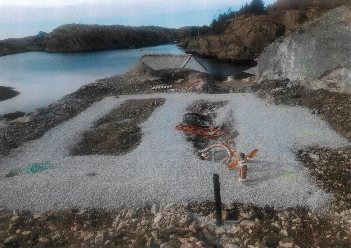 Byggeplass med grus og verktøy ved vannkanten, omringet av steiner og fjell.