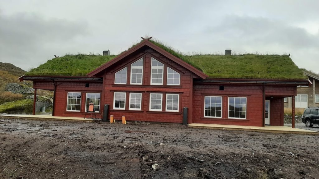Rødt hus med gressdekt tak, store vinduer og moderne design, plassert i et naturskjønt område.