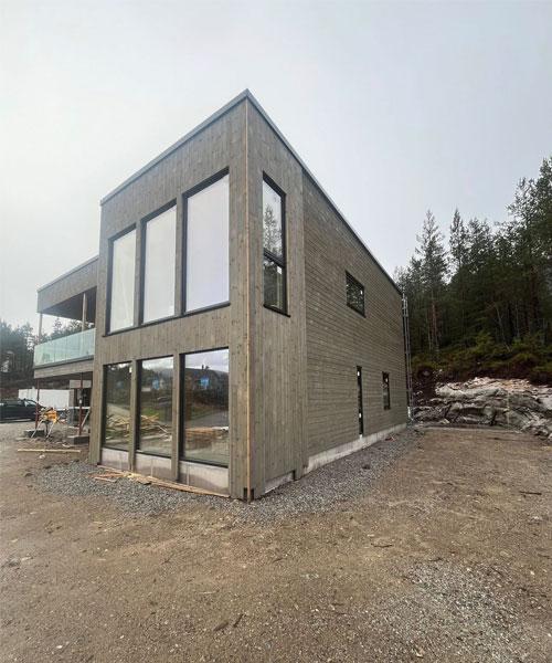 Moderne trehus med store vinduer, plassert i et skogkledd område med grusunderlag.