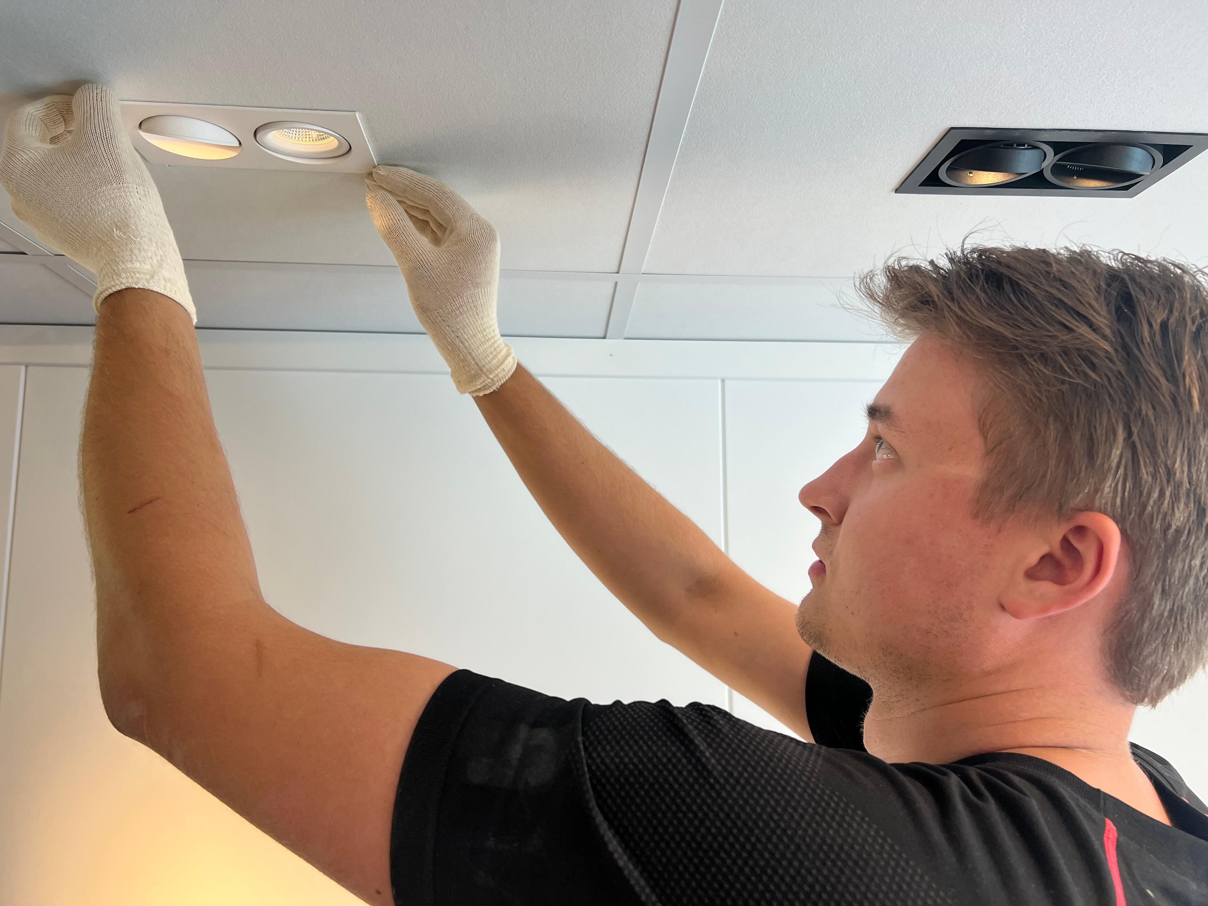 En mann med hansker skifter en taklampe i et moderne rom med hvite vegger.