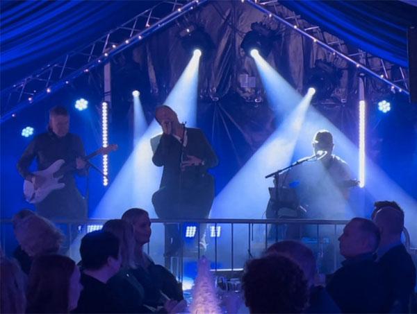 En musikkonsert med en sanger og to musikere på scenen, opplyst av blå lys, foran et publikum.