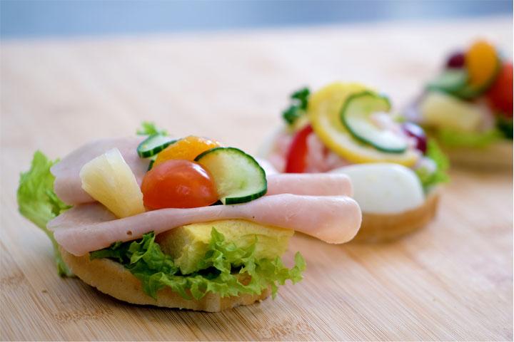Tre smørbrød med salat, skinke, agurk, tomater og frukt på en treplate.