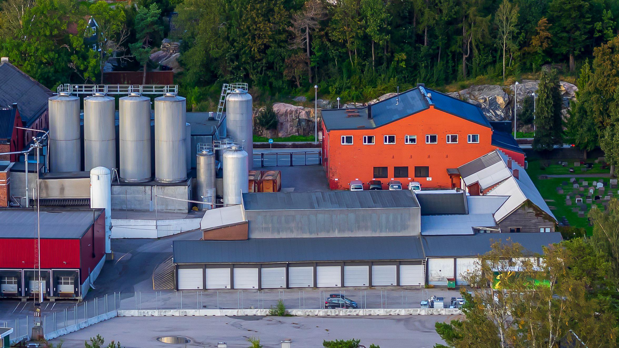 Fabrikkbygning med sølvfargede tanker og rød murvegg, omgitt av trær og parkeringsplasser.