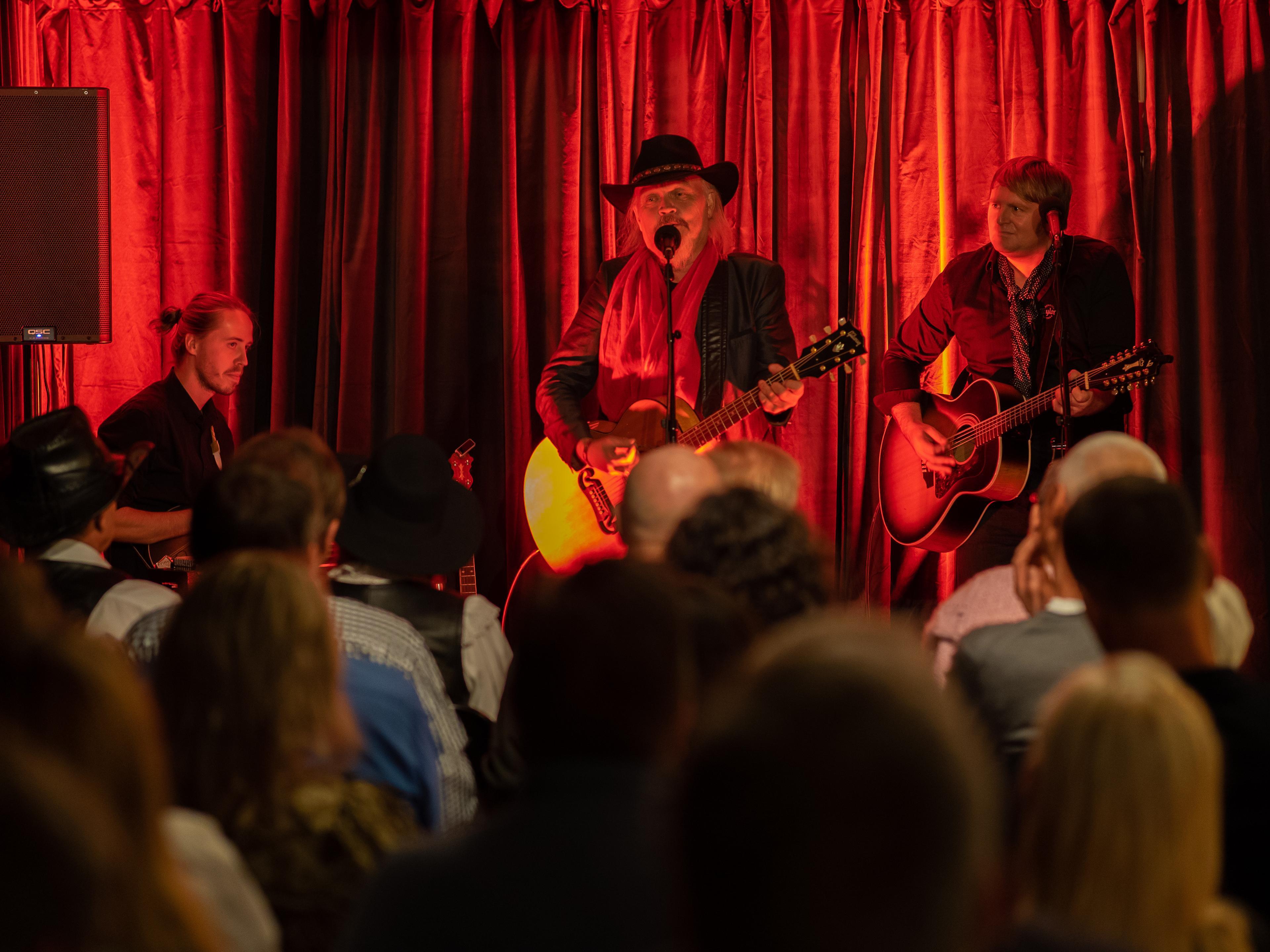 En musikkonsert med en sanger og to gitarister på scenen, med rødt lys og et publikum som ser på.