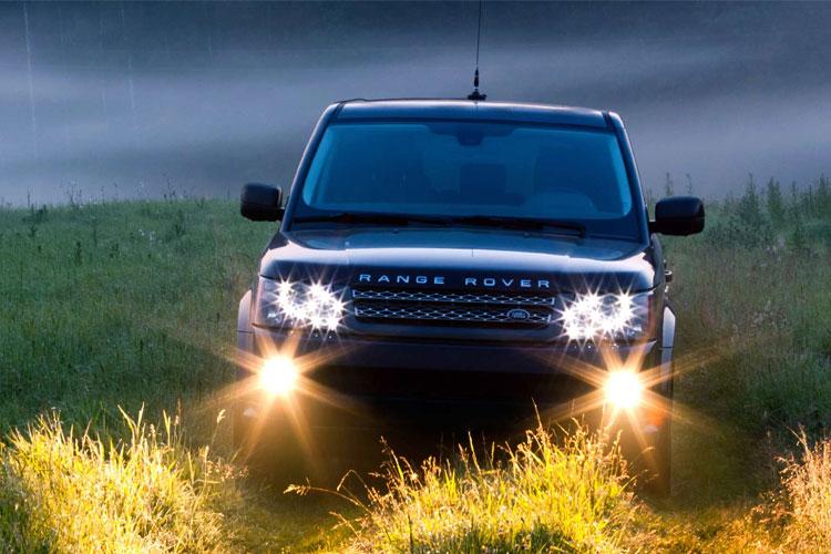 En svart Range Rover med lysene på, kjører gjennom en grønn eng i tåke.