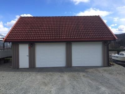 En garasje med to hvite porter og rød takstein, plassert på grusunderlag under en blå himmel.