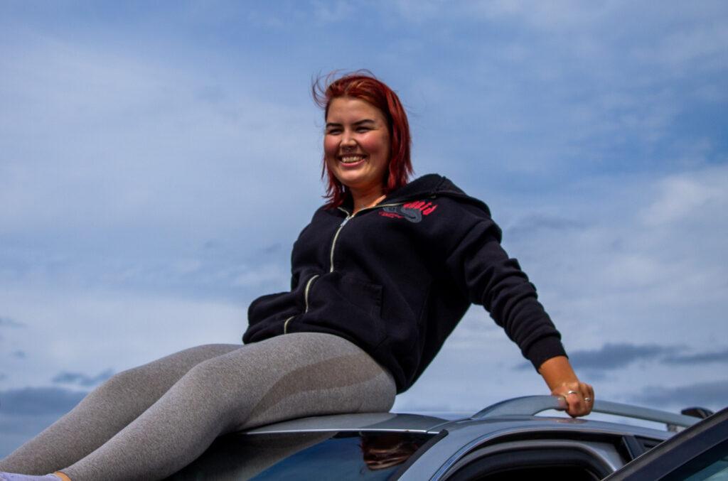 Ung kvinne med rødt hår sitter på taket av en bil under en blå himmel. Hun smiler og har på seg en svart hettegenser.