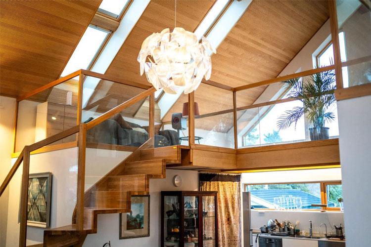 Moderne stue med tretrapp, glassrekkverk og stor lampe, samt planter i et lyst rom med skråtak.
