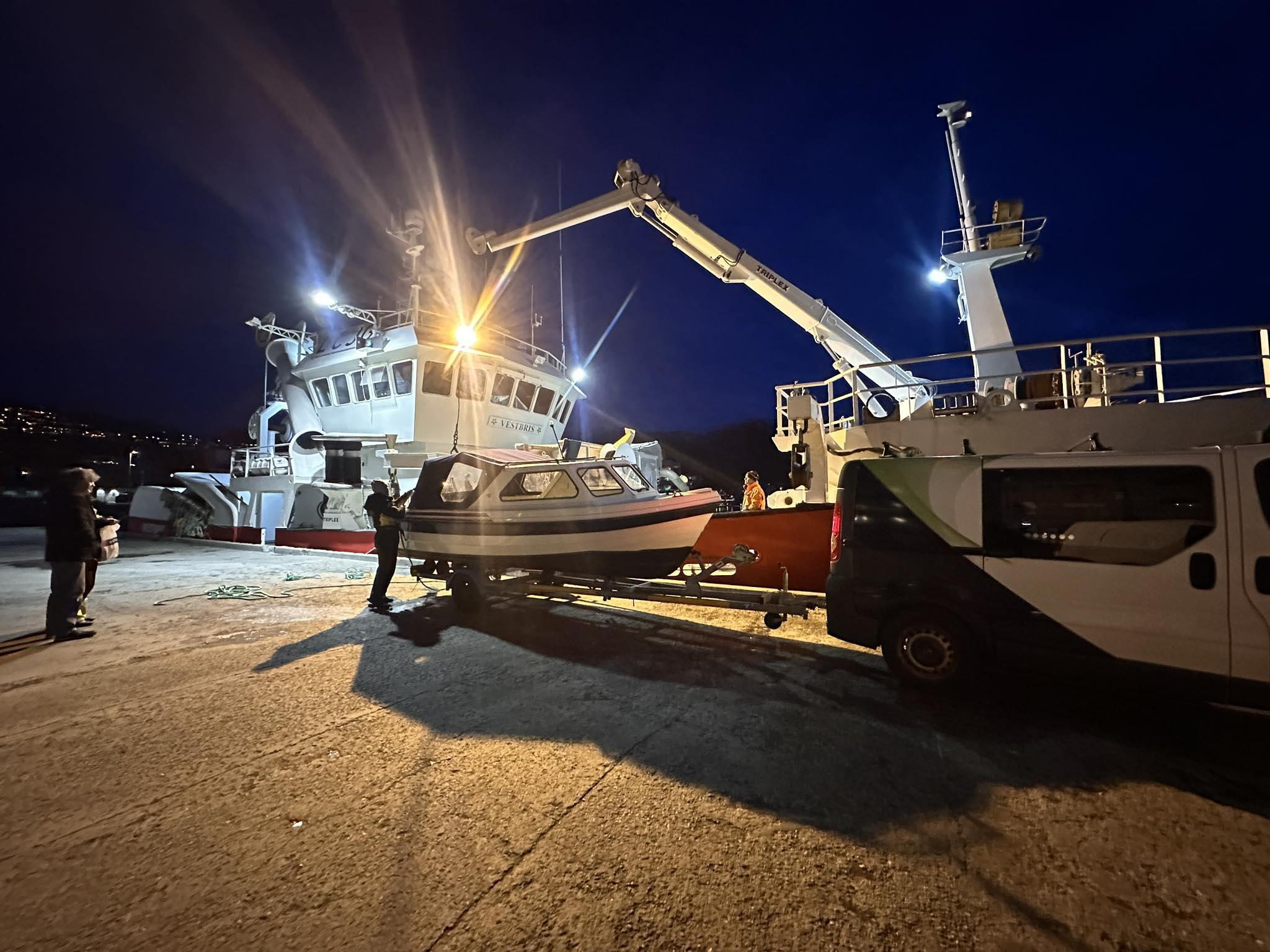 Fiskebåt blir lastet med en liten båt ved kai om natten, opplyst av sterke lys.