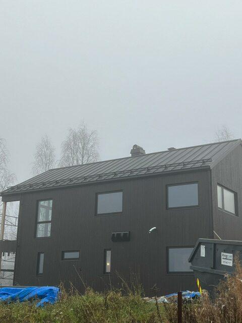 Moderne svart hus med metalltak, omgitt av tåke og trær, med en søppeldunk i forgrunnen.