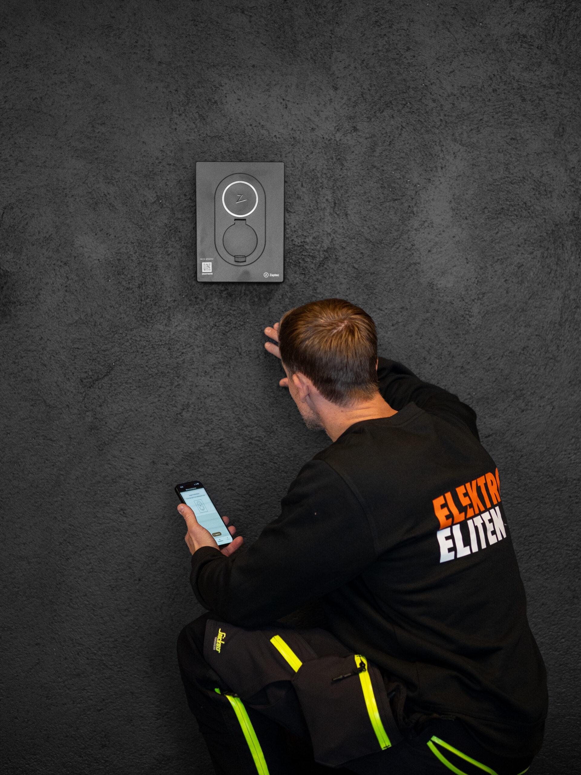 En elektriker i arbeid installerer en moderne dørklokke på en mørk vegg, mens han sjekker telefonen sin.