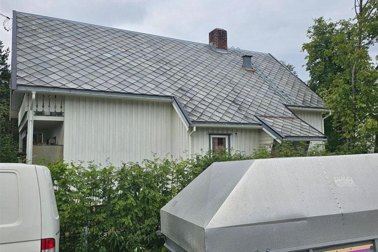 Et hvitt hus med skråtak og grå takstein, delvis skjult av busker og en søppelkasse i forgrunnen.