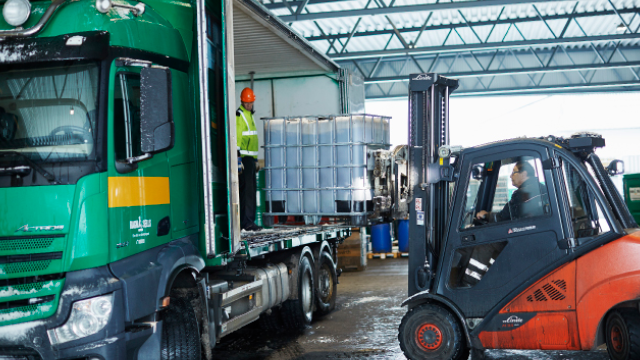 Lastebil som laster av store tanker med gaffeltruck i et lagerområde.