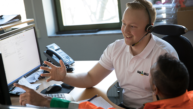En smilende mann med headset snakker med en kollega ved et skrivebord med datamaskin og telefon.