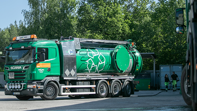 Grønn søppelbil fra Scania står parkert ved en avfallsstasjon med ansatte i arbeid i bakgrunnen.