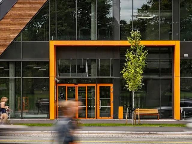 Moderne bygning med oransje inngang, store vinduer og en liten treplante foran. Folk går forbi på fortauet.