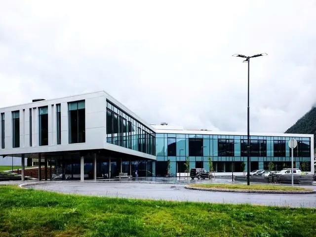 Moderne kontorbygg med store vinduer og grønt område foran, beliggende i et naturskjønt landskap.