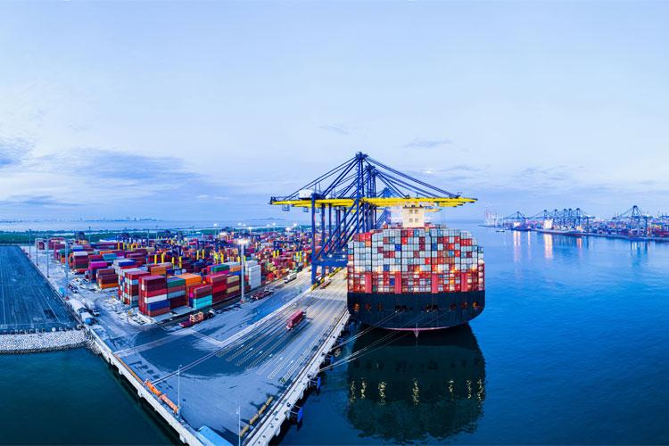 Et stort containerskip ligger ved kaien, omgitt av fargerike containere og havet under en blå himmel.