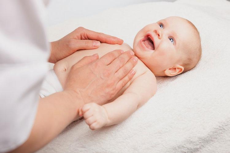 En baby ligger på et mykt underlag mens en voksen masserer brystet hennes, og hun smiler glad.