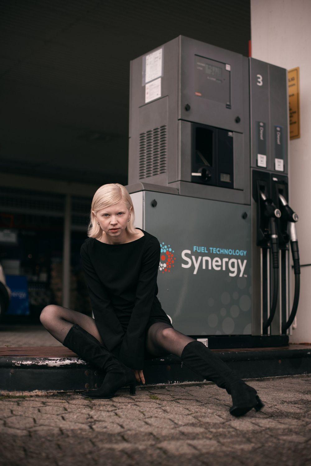 En kvinne med blondt hår sitter på bakken foran en bensinstasjon med drivstoffpumper.