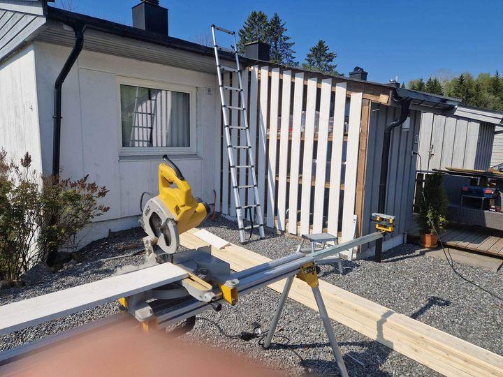 Byggeplass med en sirkelsag, stiger og en vegg med treplanker under solfylt himmel.
