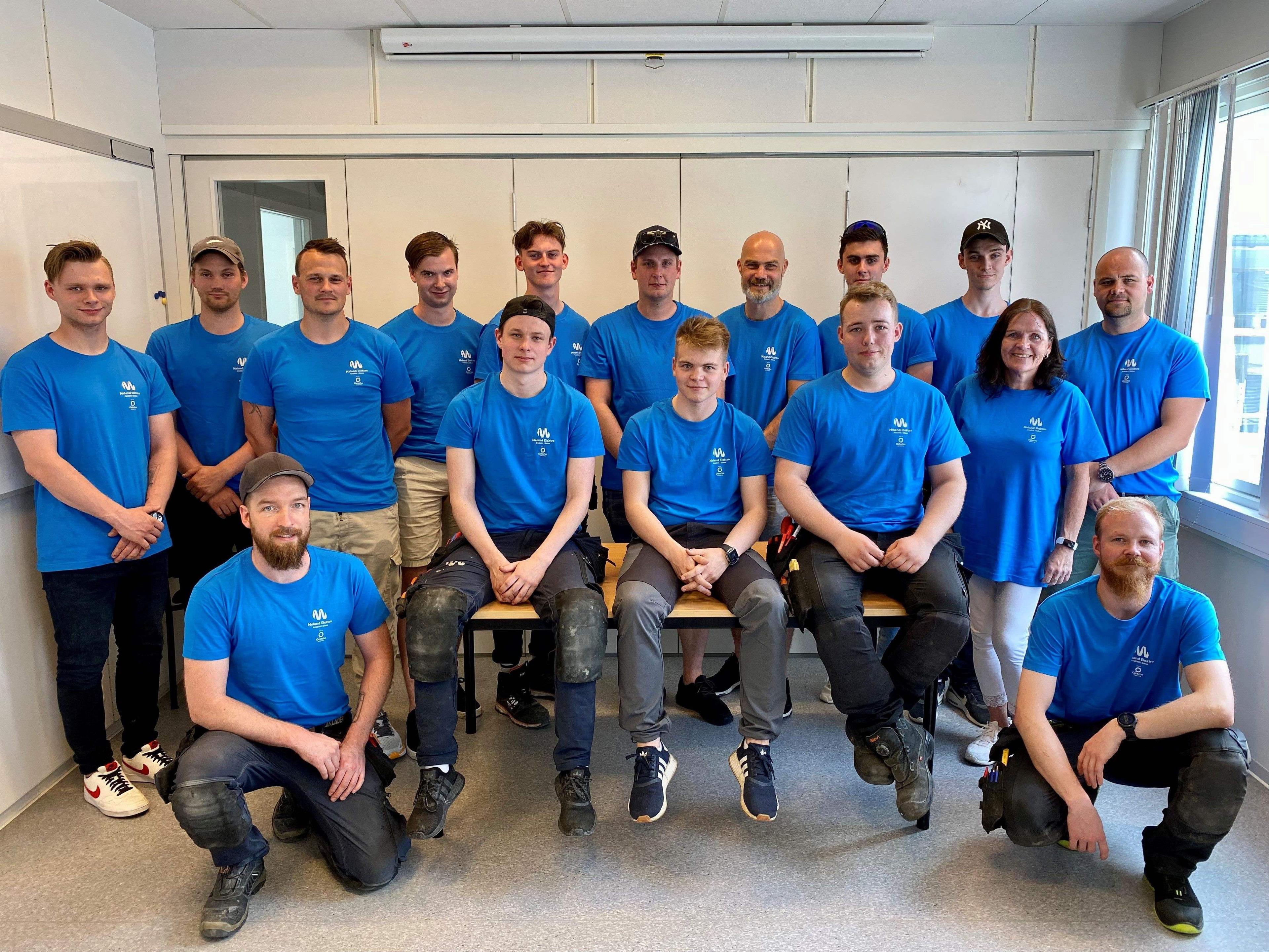 Gruppebilde av en gruppe menn og kvinner iført blå t-skjorter, samlet i et rom med vinduer og kontormøbler.