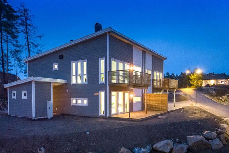 Moderne enebolig med store vinduer, balkong og belysning, beliggende i et rolig nabolag ved skumring.