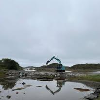 Gravemaskin arbeider i et våtmarksområde med steiner og vann under en grå himmel.