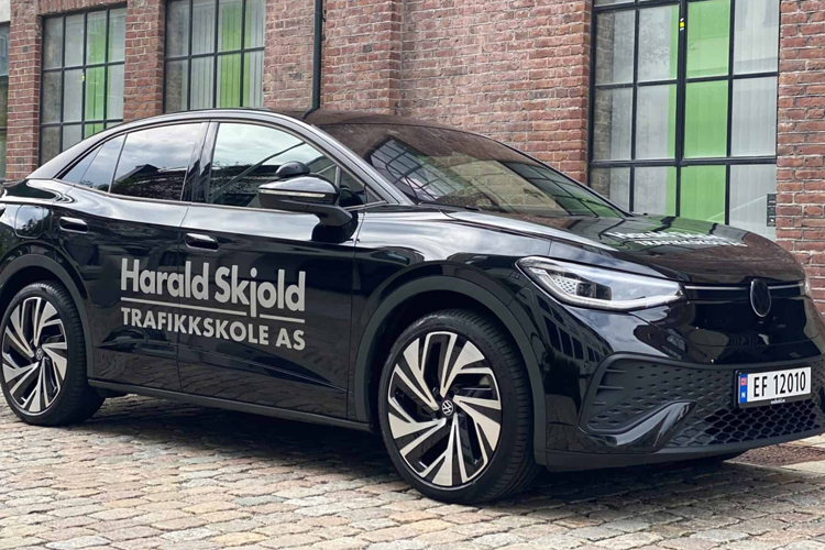 Svart elbil med teksten "Harald Skjold Trafikkskole AS" parkert foran murvegg med grønne vinduer.