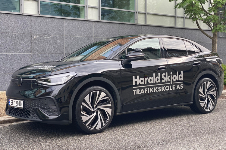 En svart bil med teksten "Harald Skjold Trafikkskole AS" parkert på en gate med bygninger i bakgrunnen.
