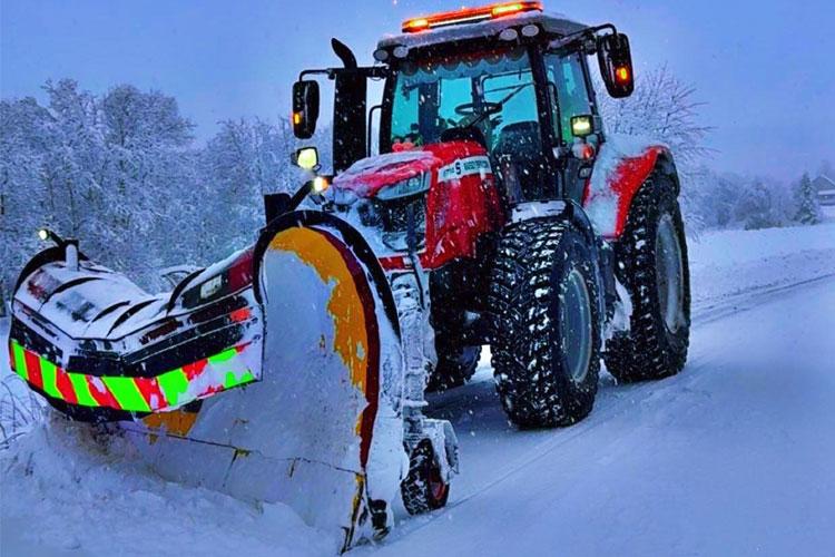 Rød traktor med snøplog som rydder snø fra en vintervei i snøfylt landskap.