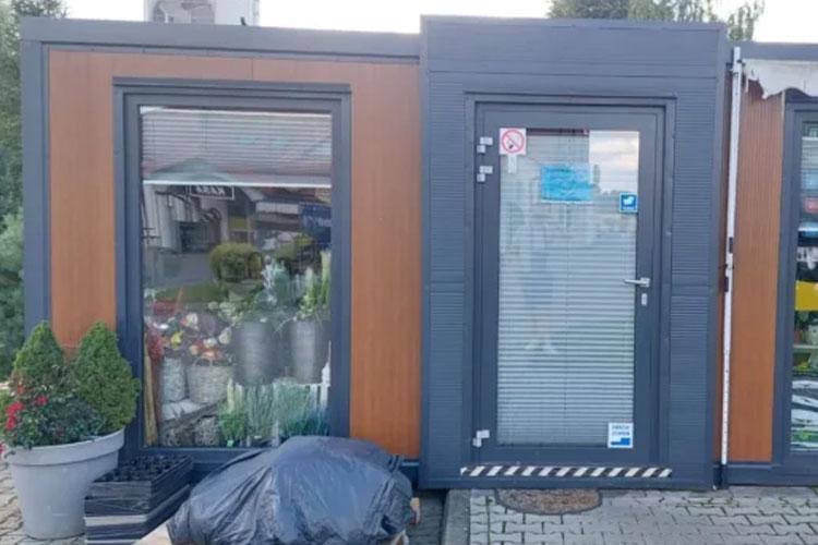 Moderne butikk med store vinduer, inngangsdør og planter utenfor, plassert på en steinlagt gårdsplass.