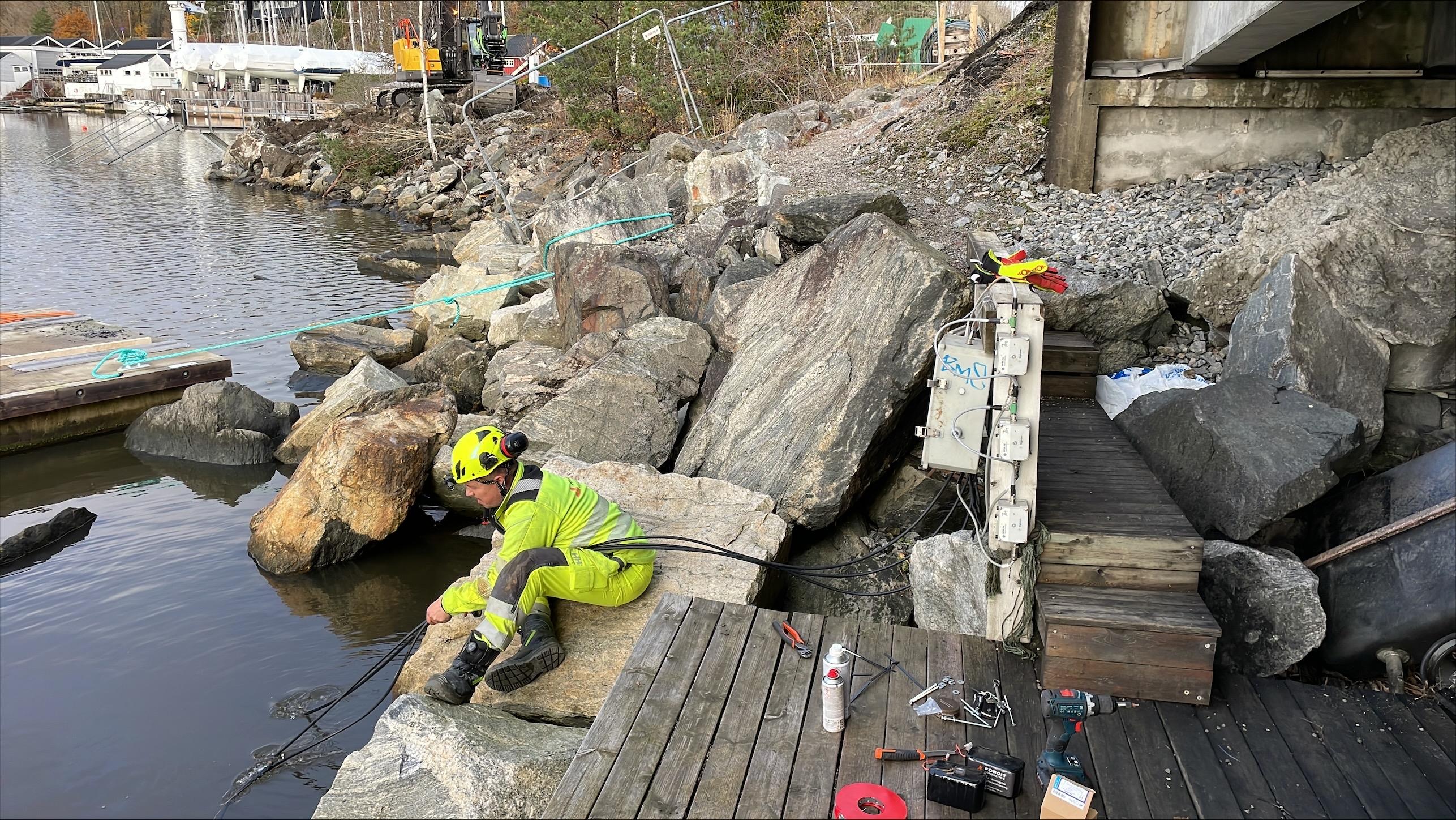 En arbeider i sikkerhetsutstyr jobber ved en brygge med steiner og verktøy langs vannkanten.