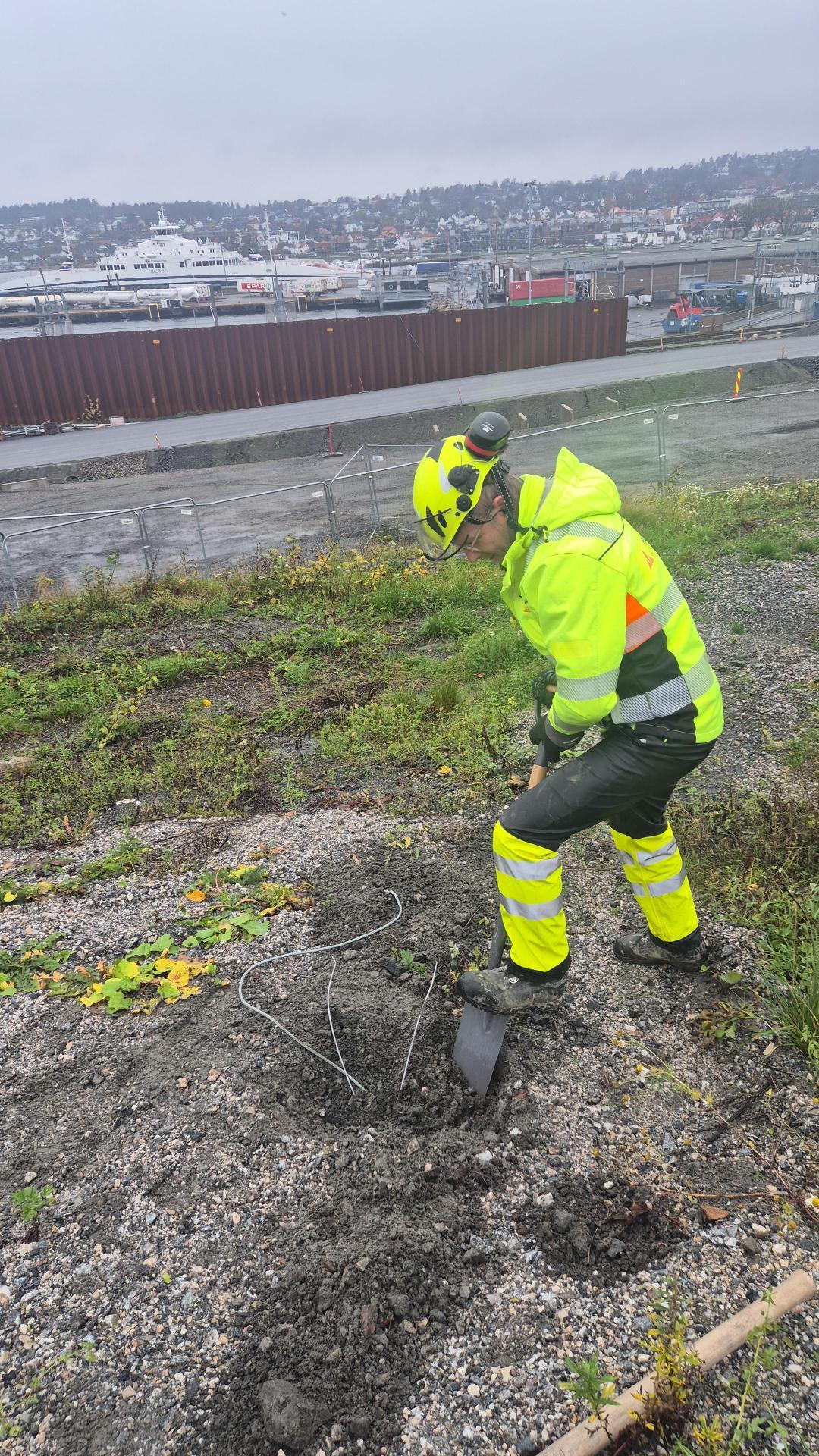 En arbeidstaker i refleksjakke graver i bakken med en spade, med en byutsikt i bakgrunnen.