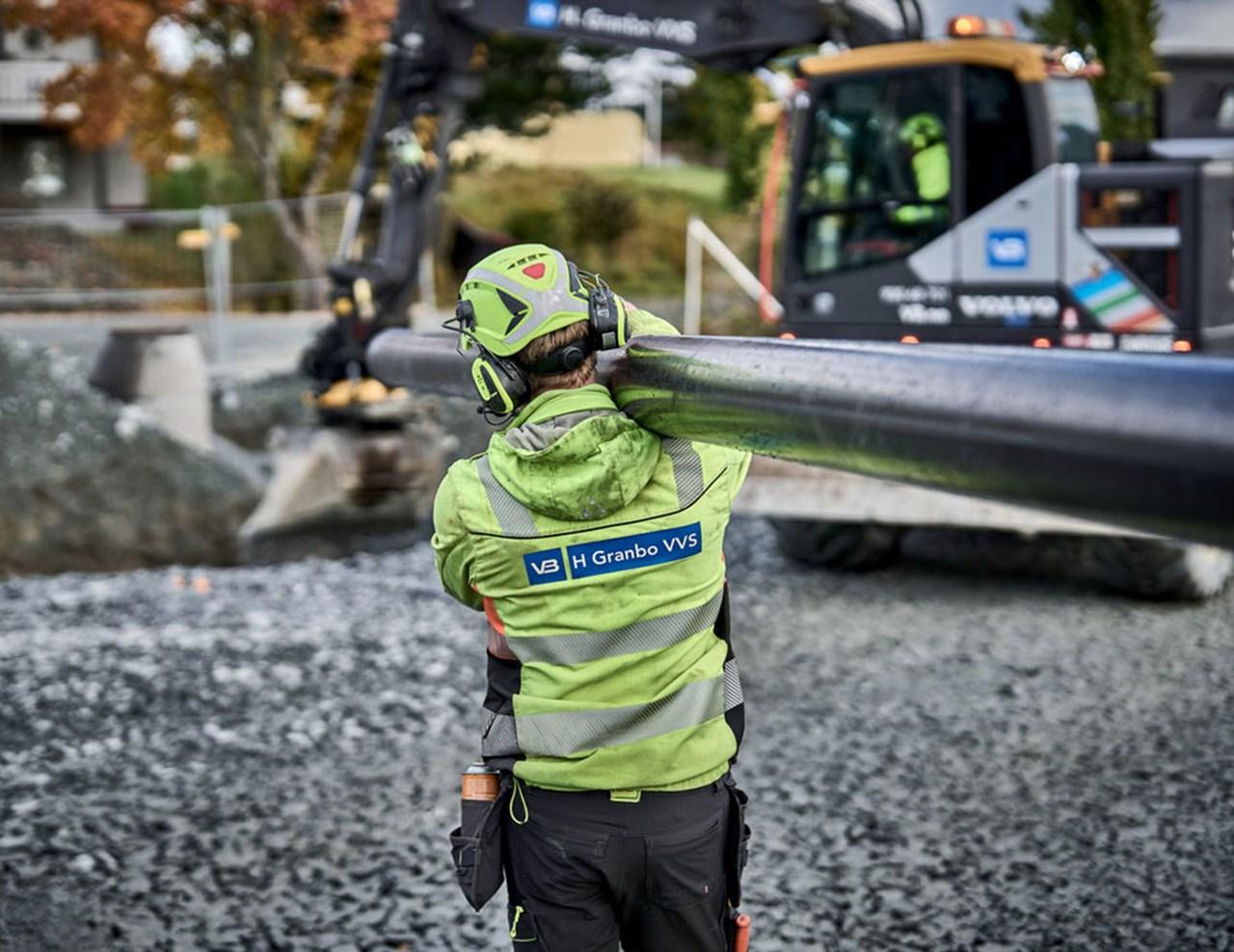 En arbeidstaker bærer en lang rørledning mens en gravemaskin jobber i bakgrunnen.