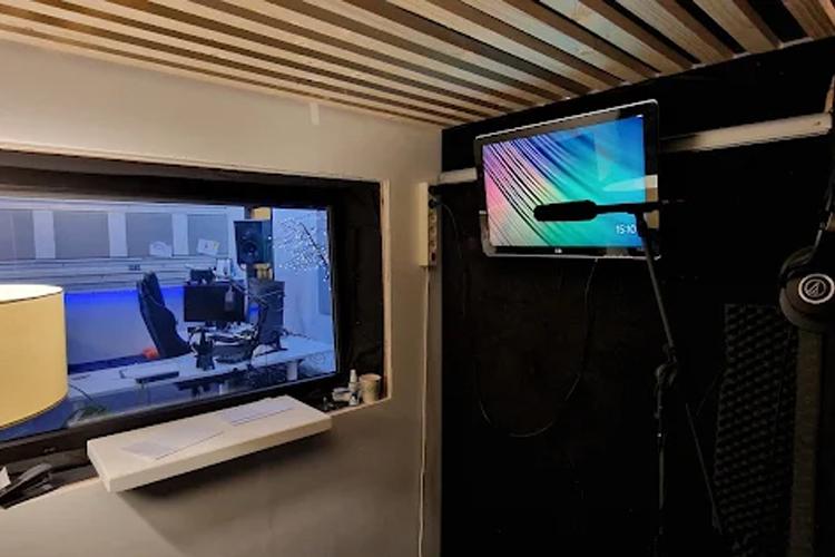 Innendørs studio med mikrofon, dataskjerm og vindu som viser et møterom.