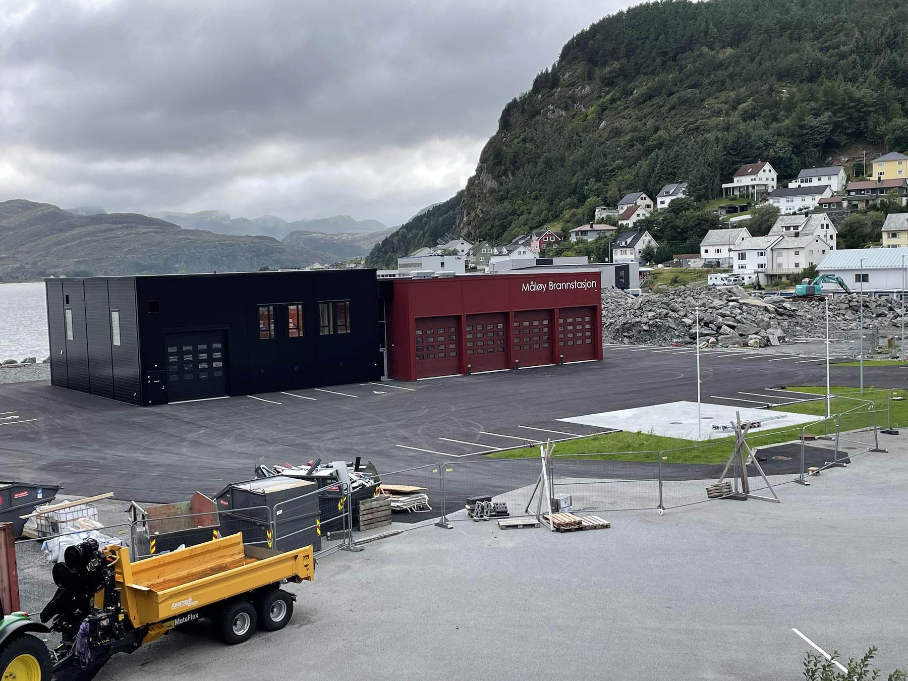 En moderne bygning med garasjeporter ved vannet, omgitt av fjell og hus i bakgrunnen.