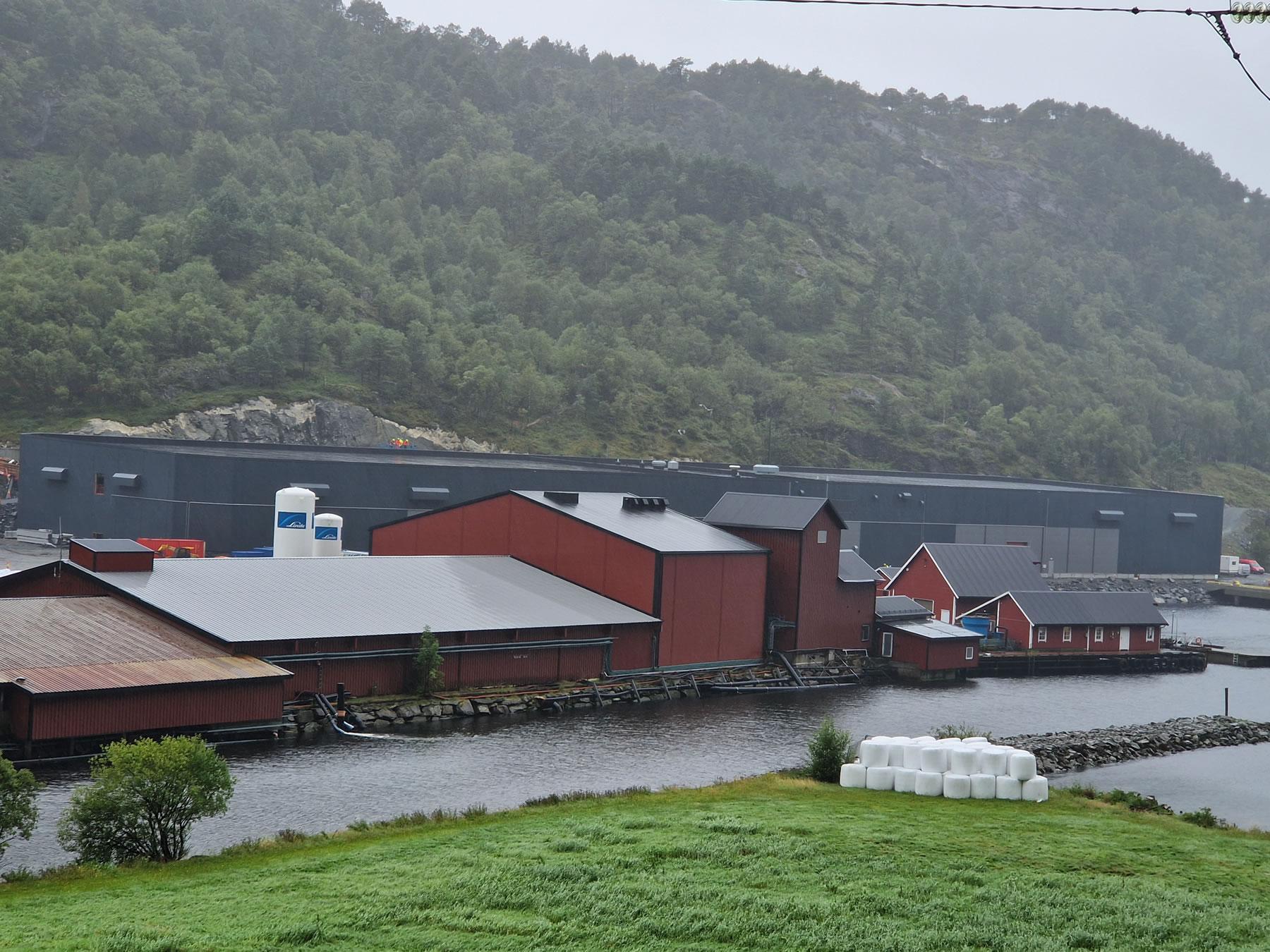 Utsikt over en rød bygning ved vannet med fjell i bakgrunnen og moderne bygninger i grått.