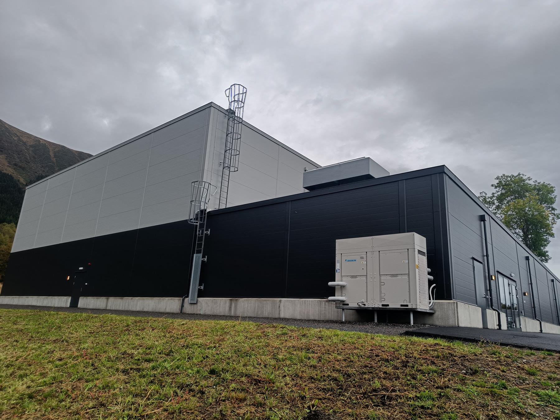 Moderne svart og grå bygning med antenne, plassert i et grønt område under en overskyet himmel.