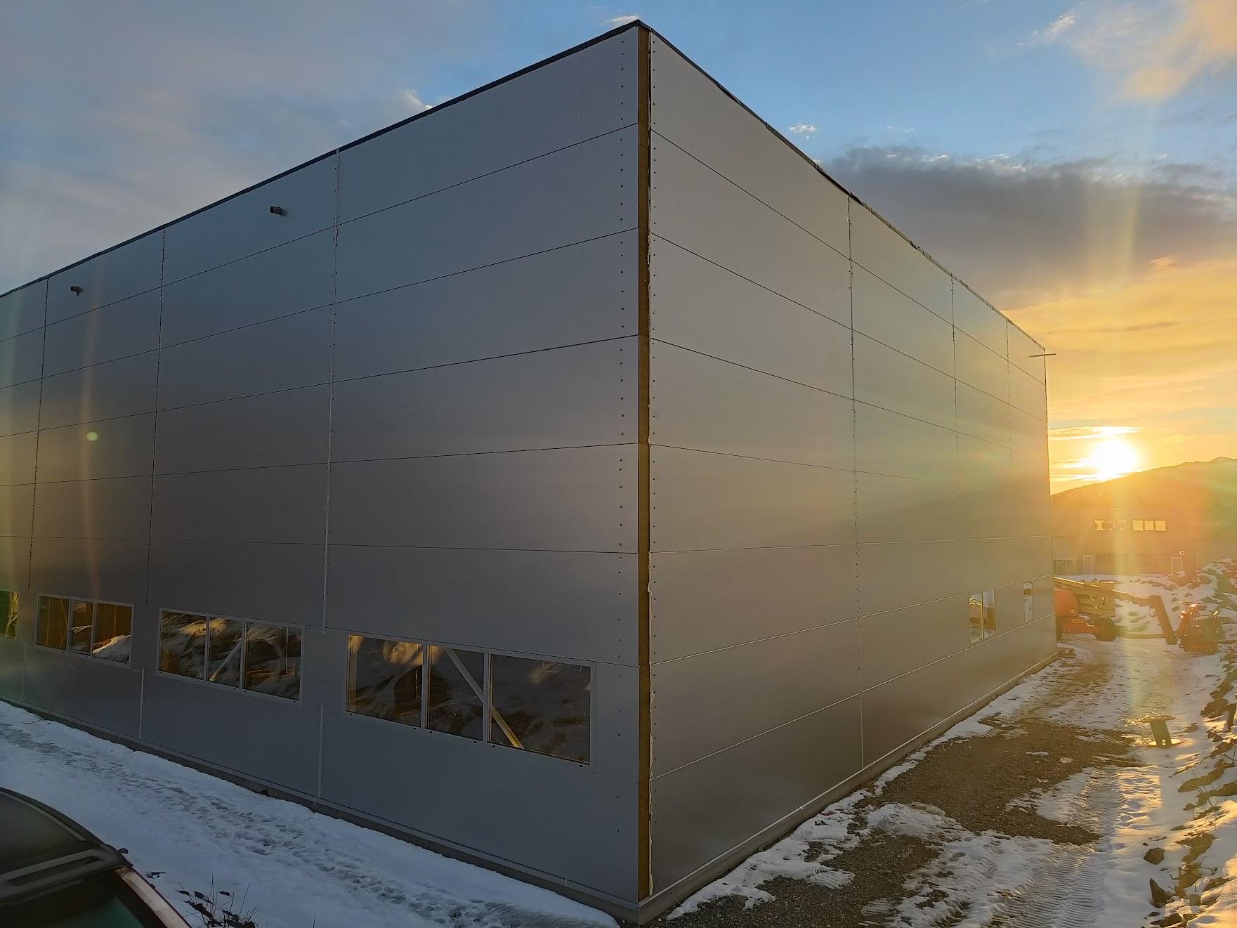 Moderne bygning med metallfasade, belyst av solnedgang, omgitt av snø og grus.