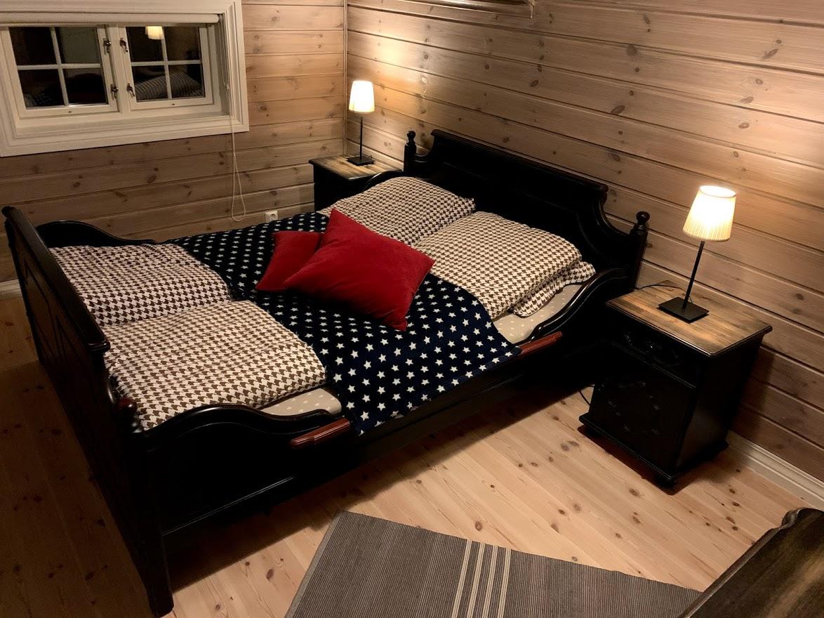 Koselig soverom med en dobbeltseng, puter, sengetøy og nattbord med lampe. Veggene er i trepanel.