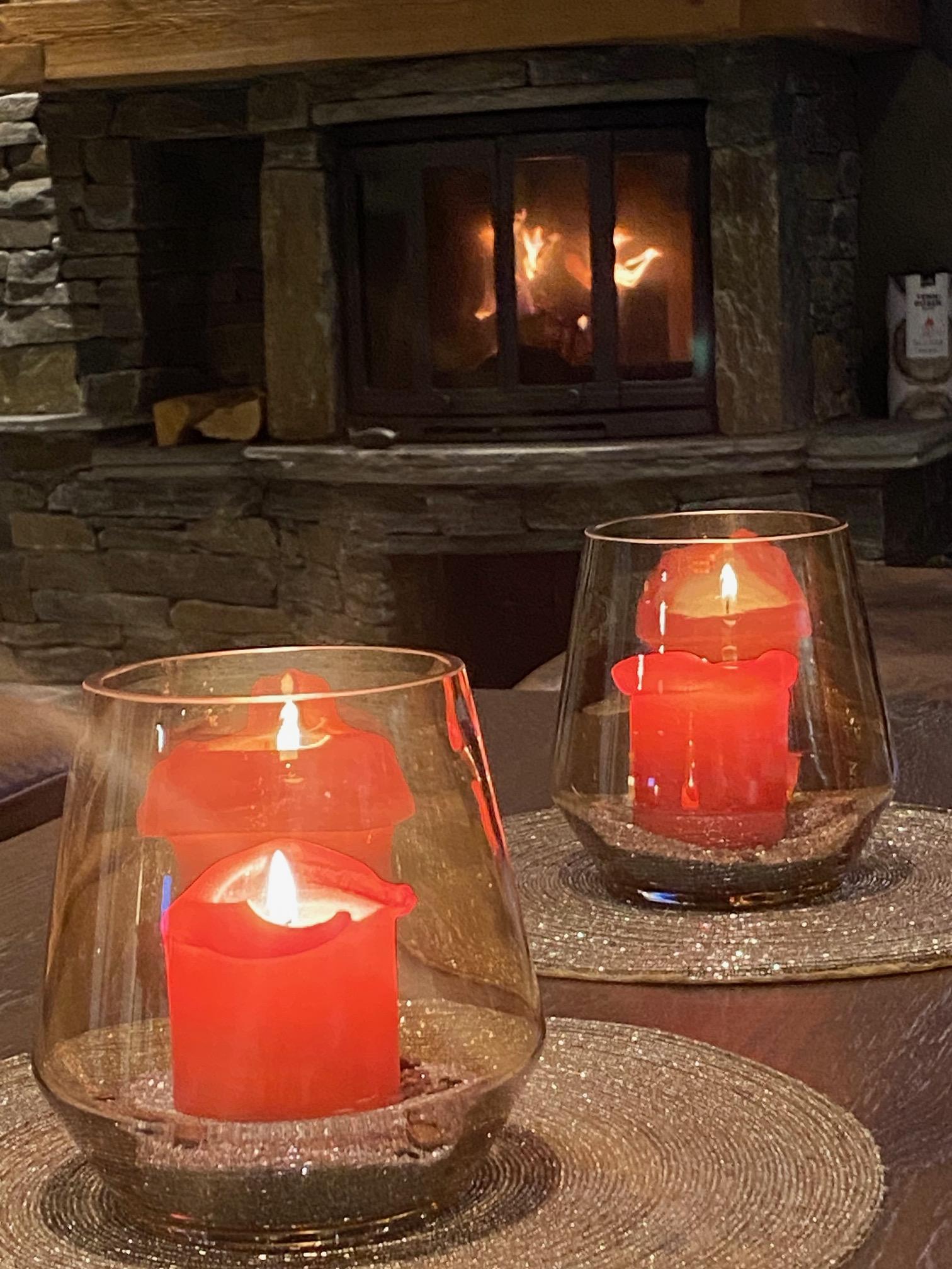 To lys med røde stearinlys i glass, plassert på et bord foran en peis med flammer.