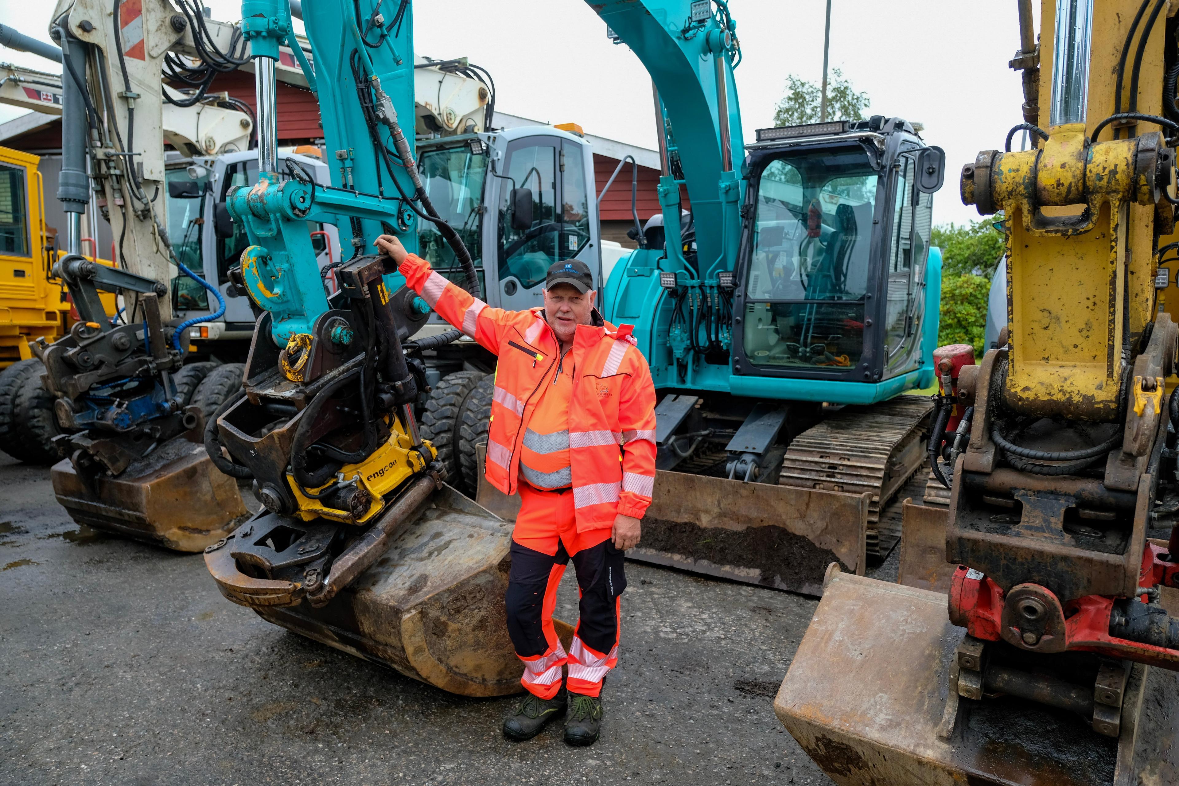 En mann i oransje arbeidsklær står ved siden av gravemaskiner på en byggeplass.