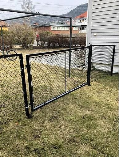 Svart gjerde med netting i en hage, med en åpen port og grønne plener i bakgrunnen.
