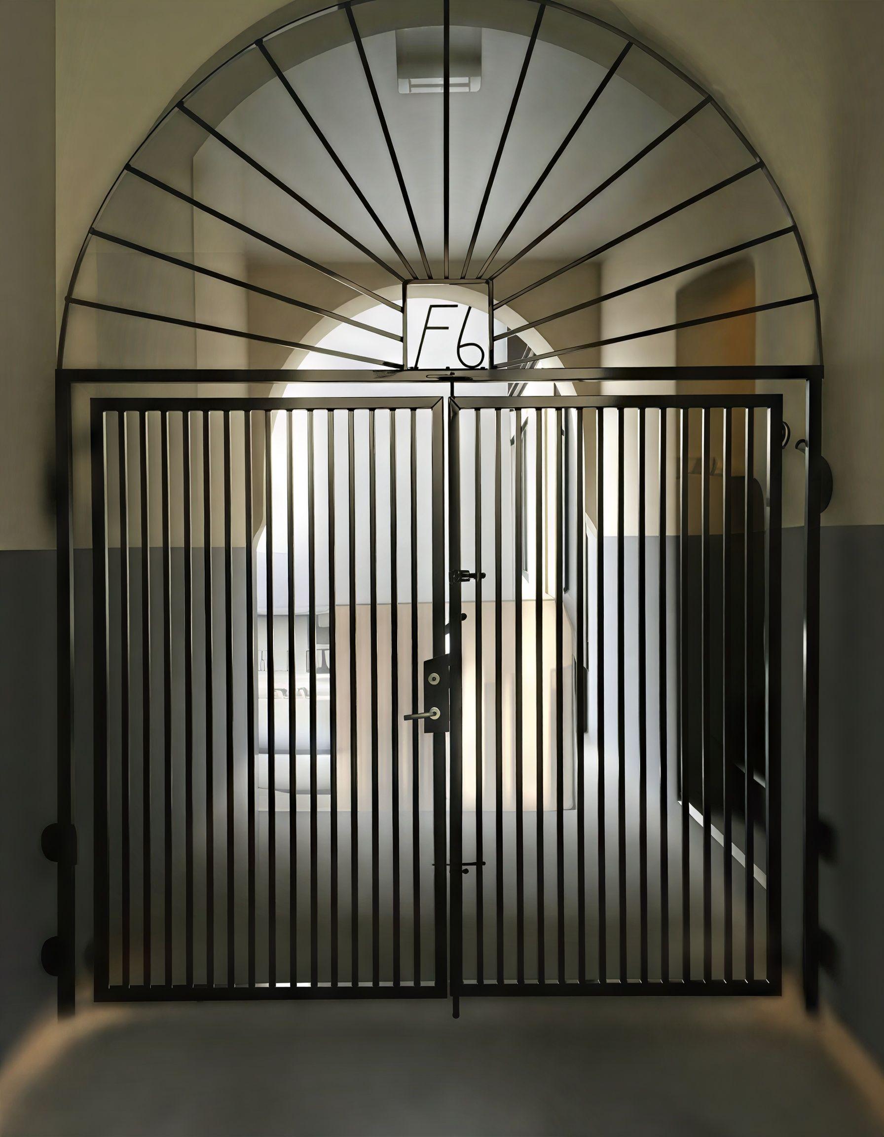 En svart jernport med buet topp, som fører inn til en gang med lys og vegger i grått.