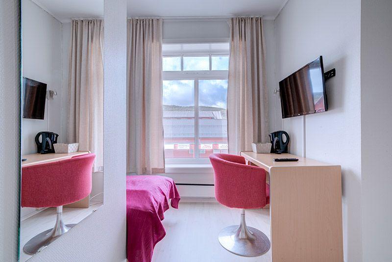 Moderne hotellrom med en enkeltseng, skrivebord, stoler, TV og vinduer med gardiner.